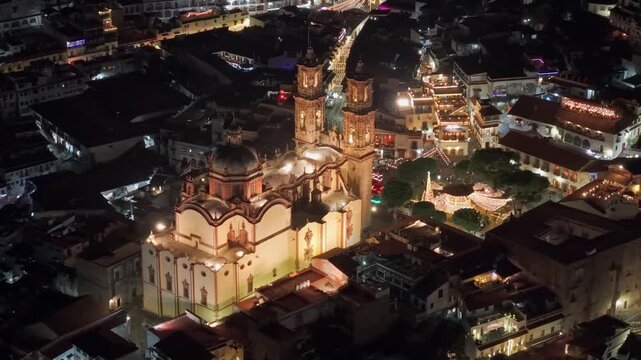 Isolated telephoto aerial view of the Santa Prisca temple illuminated at night with lights in the square and white colonial houses around it, Taxco, Guerrero, Mexico