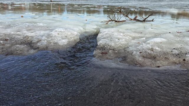 Rapid snowmelt cuts a dark channel through dirty spring ice, as leftover snow softens and rushes into a narrow stream at ground level.
