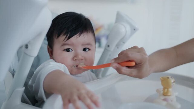 4K slow motion footage of an Asian mother feeding her six month old baby boy with a spoon. Tender parenting moment showing care, bonding, and infant nutrition in a warm home environment.	
