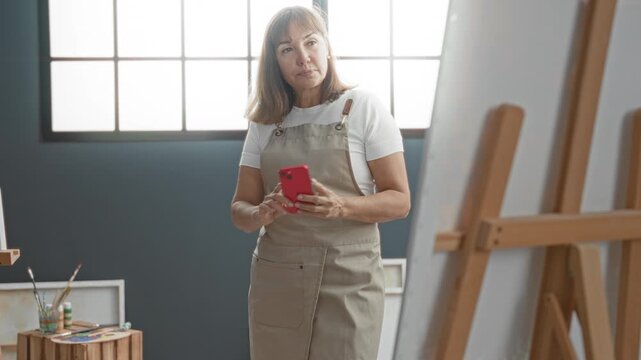 Middleaged woman painter in apron holds smartphone and taps screen in studio near easel and paint jars; focused creativity.