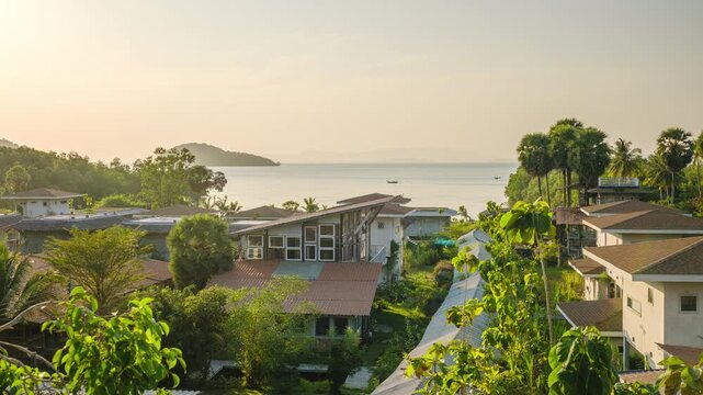 Beautiful sunset timelapse transitioning to night over a coastal resort in Thailand. Features changing sky colors, village rooftops, lush greenery, and the ocean bay under a dark starry sky.