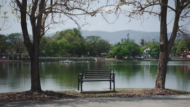 Static shot of an empty metal park bench positioned between two trees, facing a serene lake with distant mountains in the background.