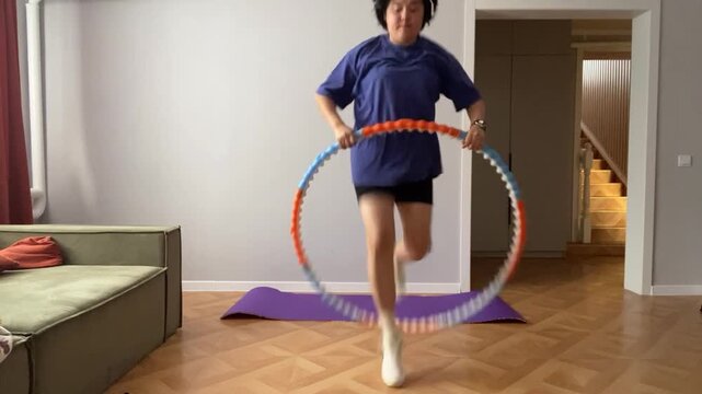 A middle-aged Asian man exercises with a hula hoop in a bright living room. He wears a blue t-shirt and black shorts, showcasing a fitness routine.