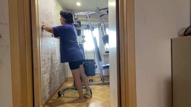 A young Asian woman stands in a home gym, adjusting a wall-mounted exercise chart. Fitness equipment is visible in the background.