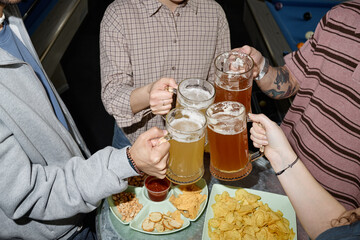 Group of young adults clinking beer mugs over table with snacks, celebrating together in casual...