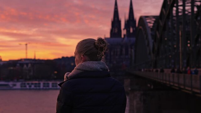 A woman seated on the Rhine River promenade, she looks across the river. She is the central subject, framed by the massive architecture of the Cologne Cathedral and the Hohenzollern Bridge.