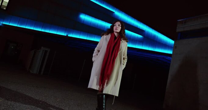 Wide-angle cinematic shot of a woman walking at night beneath blue neon lighting, wearing a coat and red scarf in a modern urban setting. Concept for nightlife, independence, street fashion, city.