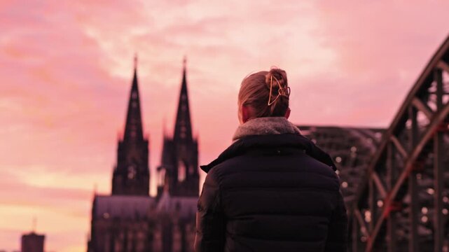 Woman admiring the iconic Cologne skyline from the Rheinboulevard in Deutz. The Hohenzollern Bridge and the Cologne Cathedral against a fiery sunset sky.