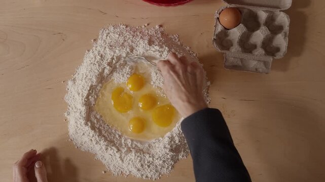 Overhead shot of a Romagna cook mixing eggs and flour with a fork to prepare fresh cappelletti dough. Traditional Italian pasta-making using raw ingredients on a wooden table.