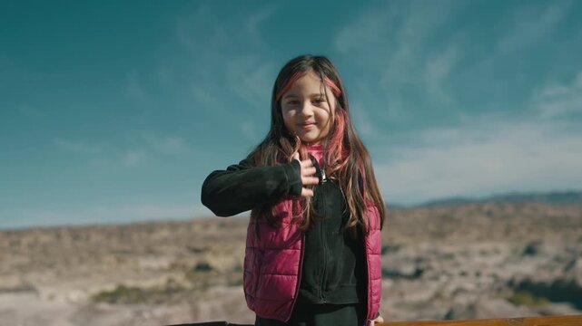 Closeup portrait of a young girl in a black top and red vest waving her hand and smiling at the camera, set against blurred desert background of Ischigualasto Provincial Park.
