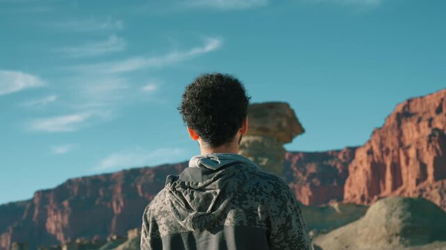 Rear view of a male tourist in a camo hoodie standing still and admiring El Hongo rock formation in Ischigualasto, San Juan, contrasting unique geological shapes against deep blue sky.