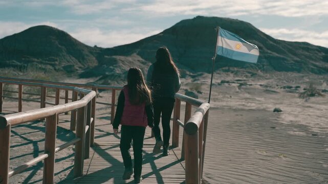 Still clip of mother and young daughter walking along tourist wooden pathway at Ischigualasto (Valle de la Luna) in San Juan, Argentina. Scene features flag, desert hills, and desaturated color tone.
