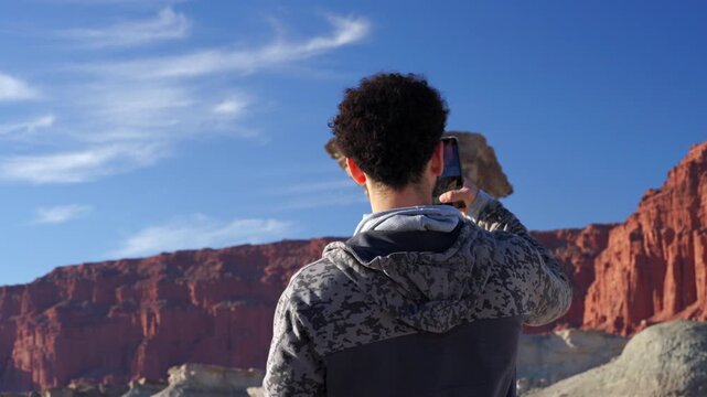 Still shot of male tourist in a camouflage hoodie using his smartphone to record the famous El Hongo rock formation at Ischigualasto, with grey rock, orange hills, and blue sky background.