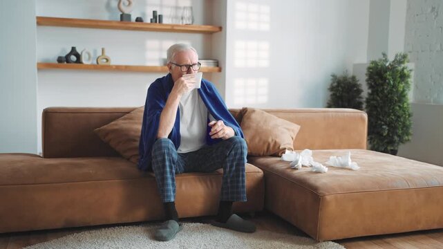 Senior man sitting on couch in cozy living room, covid pandemic. Elderly male person suffering fever, runny nose and cold, grippe epidemic in winter or autumn season, sick male person