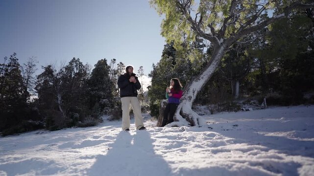 Mother and daughter share a sunny meal, eating a sandwich, in mixed forest clearing, seated beneath mature native Nothofagus tree, the snowy ground and alpine ecosystem, wilderness family recreation.