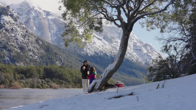 Mother and daughter pause their mountain adventure to share sandwich meal beneath a towering native tree, snow-covered landscape and distant andean peaks framing moment of family bonding and leisure.