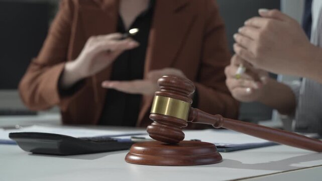 Close-up of judge gavel on desk during legal consultation, with lawyers or clients discussing paperwork in blurred background, courtroom or law office setting.