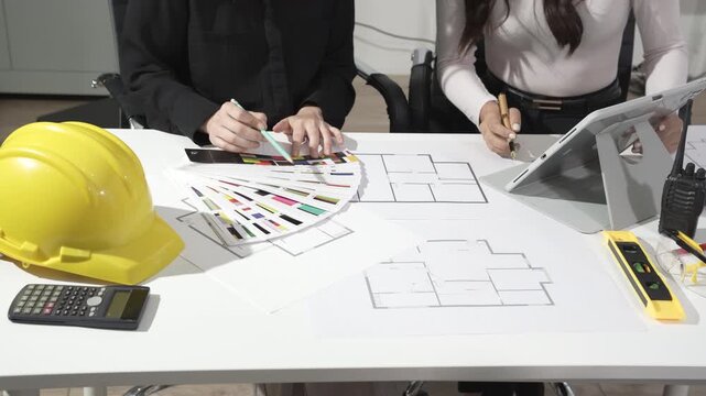 Designers and engineers collaborating at desk with architectural floor plans, color swatches, tablet, hard hat, calculator and measuring tools, planning construction or interior design project.