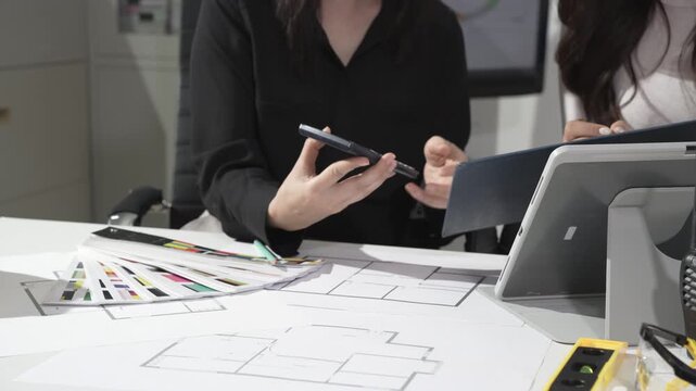 Designers reviewing architectural floor plans at desk, discussing with smartphone and tablet, color swatches and tools in modern office meeting.