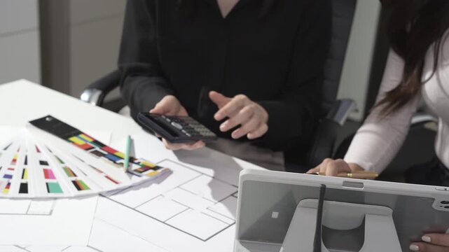 Two colleagues collaborate at a desk with color swatches, design layouts and a calculator, planning a project in a modern office setting.