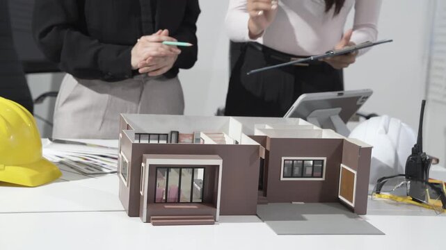 Architects review scale model of modern house on desk with hard hats, plans, tablet and clipboard in office meeting, real estate development and construction planning concept.