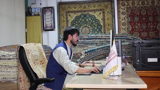 A man in Kabul city is selling rugs in large quantities in carpet shop behind computer screen. many carpets on background, Afghanistan economy and modern office work meets ancient culture concept