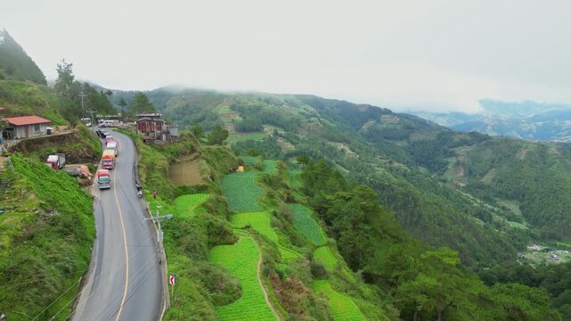 Side-gliding aerial tracking parallel to the mountain highway, showcasing vibrant tiered rice terraces on the slopes of Buguias, Benguet, Philippines.
