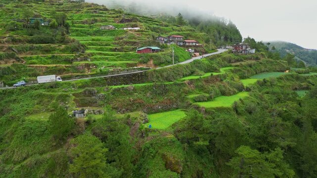 Dynamic right push aerial approaching the tiered rice terraces and the winding mountain highway under misty skies in Buguias, Benguet, Philippines.
