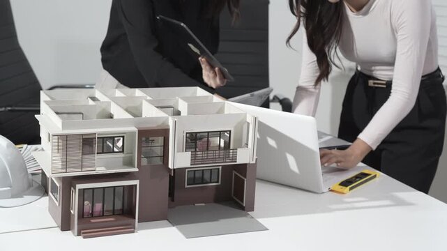 Architects review a modern house scale model on a desk, collaborating with laptop, blueprints and tools in an office setting.