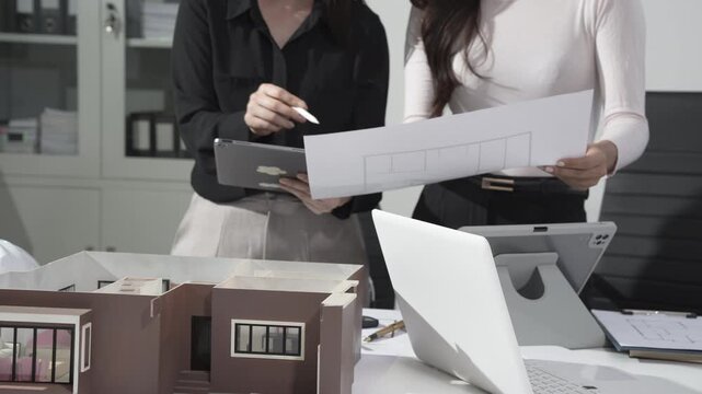 Architects reviewing floor plan with scale model on office desk, laptop and tablet, modern interior design workspace, project planning and collaboration
