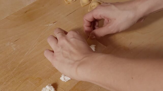 Tight view of hands folding a ricotta-filled square into cappelletti on a wooden surface in soft lighting.