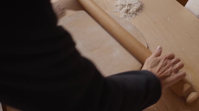 Senior hands using a wooden rolling pin to flatten dough on a floured surface, preparing homemade pasta or pastry.