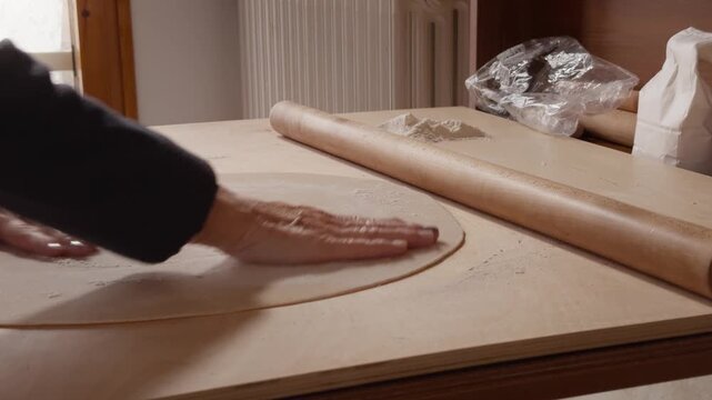 Close-up of hands using a wooden rolling pin to flatten pasta dough near a small pile of flour on a wooden table.