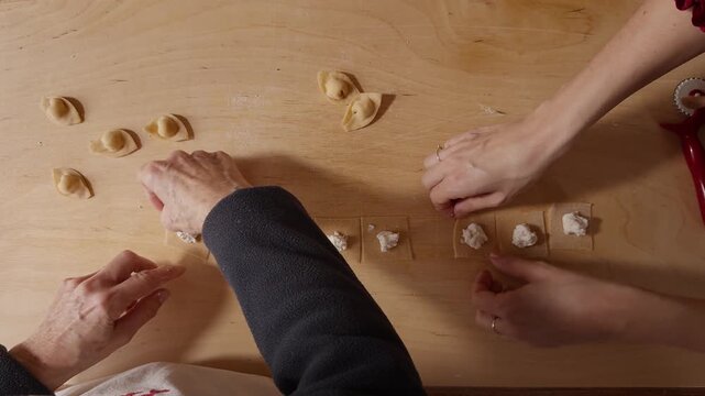 Top-down view of hands folding small pasta squares filled with ricotta on a wooden table in warm kitchen light.