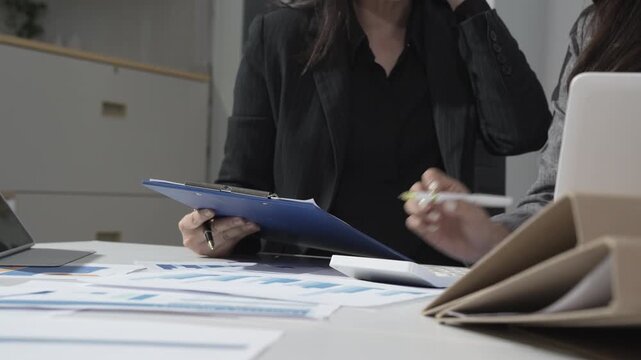 Business meeting scene with two colleagues at office desk reviewing paperwork, charts and documents; one holds clipboard and gestures while laptop and files sit on table.