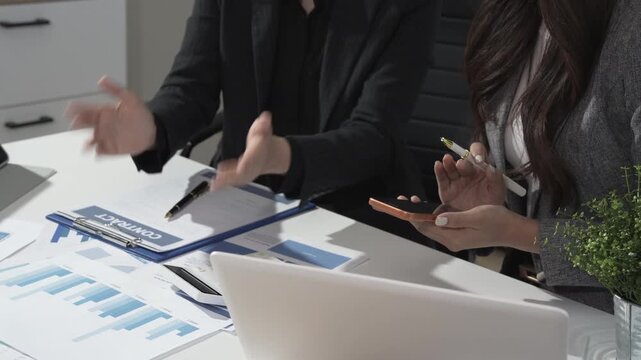 Business meeting at modern office desk with two professionals reviewing contract, charts and laptop, hands pointing and holding pen, teamwork and financial planning concept.