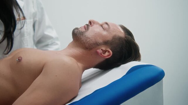 Bearded man lies calmly on a treatment table beside a therapist in a bright clinical room.