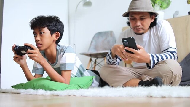 Son focuses on video game while father relaxes drinking coffee and checking smartphone at home