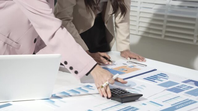 Business colleagues analyzing financial reports on desk with laptop and calculator in modern office, pointing at charts and graphs during meeting.