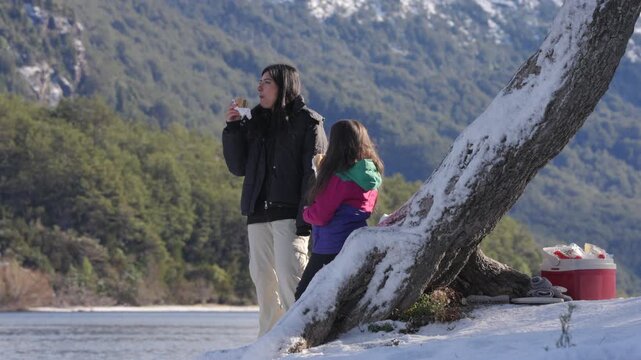 Mother and daughter eat sandwiches together beside a snowy riverbank in Patagonia, framed by forested mountains, winter light, calm water, and a quiet outdoor moment of happiness and family bonding.