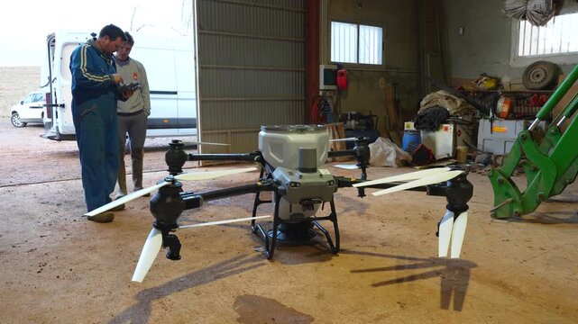 Experienced farmer instructs a younger man inside a barn on configuring an agricultural drone using the remote