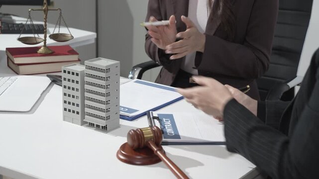 Business and legal meeting at desk with gavel, scale of justice, documents and miniature building model, suggesting property law, real estate dispute or corporate compliance.