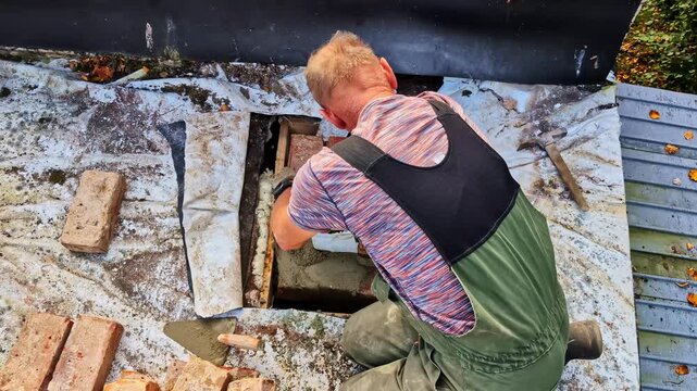 Man kneeling on a roof, performing renovation work, applying mortar to a chimney base