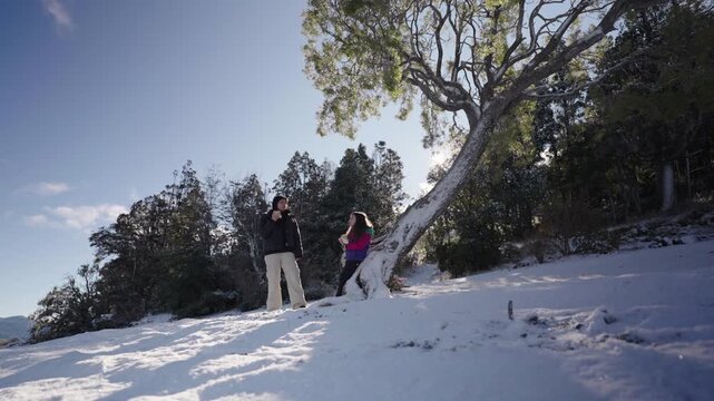 Mother and daughter stand beside a snow covered tree in a winter forest in Patagonia, sharing food together during a quiet pause surrounded by trees and mountain terrain.