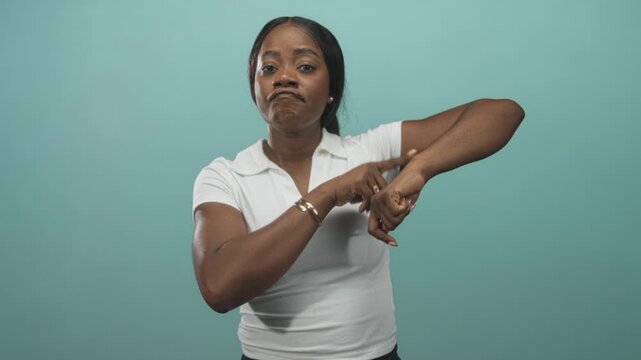 Woman points finger to wrist in studio wearing white polo, checking time with a frown and impatient pose; impatience urgency.