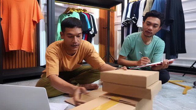 Small business partners sit on floor of apparel studio stacking shipping boxes and recording inventory on clipboard
