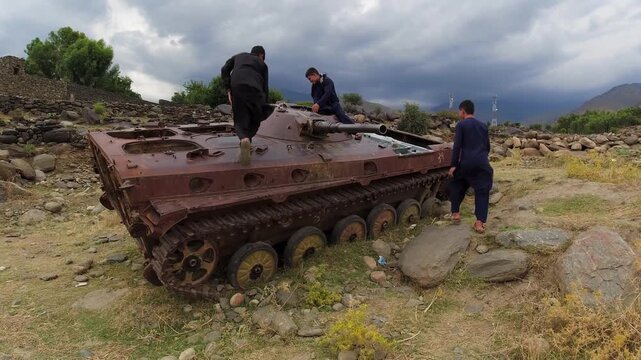 Kids climb and play on a destroyed rusty old military tank Soviet T62 in Afghanistan, Soviet American war in arid mountain landscape, Khogyani. Resilience, marks of war torn country, taliban, Kabul