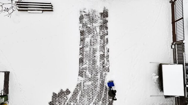 Top-down drone view of a man shoveling snow off a cobblestone walkway, revealing dark paving against a white courtyard with footprints and strong copy space.