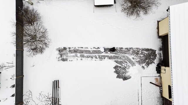 Aerial top-down static shot of a person clearing deep snow from a dark stone courtyard, revealing rhythmic pavement patterns in a minimalist winter setting,