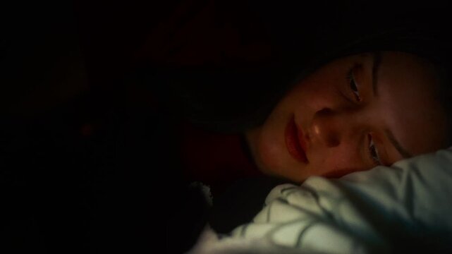 Thoughtful young woman lying in bed at night, scrolling on social media with a soft screen glow on her face, cinematic low light aesthetic.